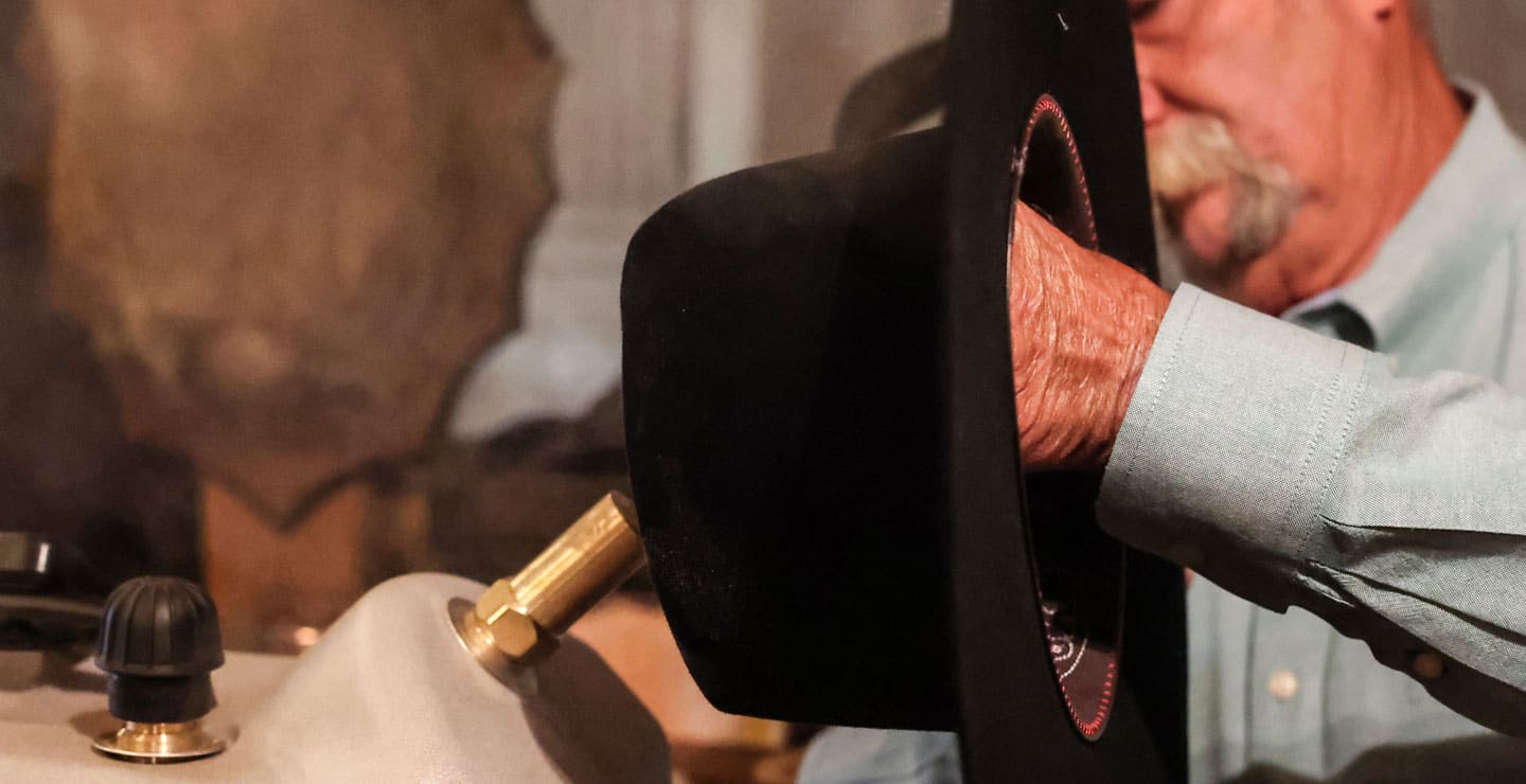An up-close photo of a cowboy with a gray mustache holding an unshaped black hat and placing it on the steamer.